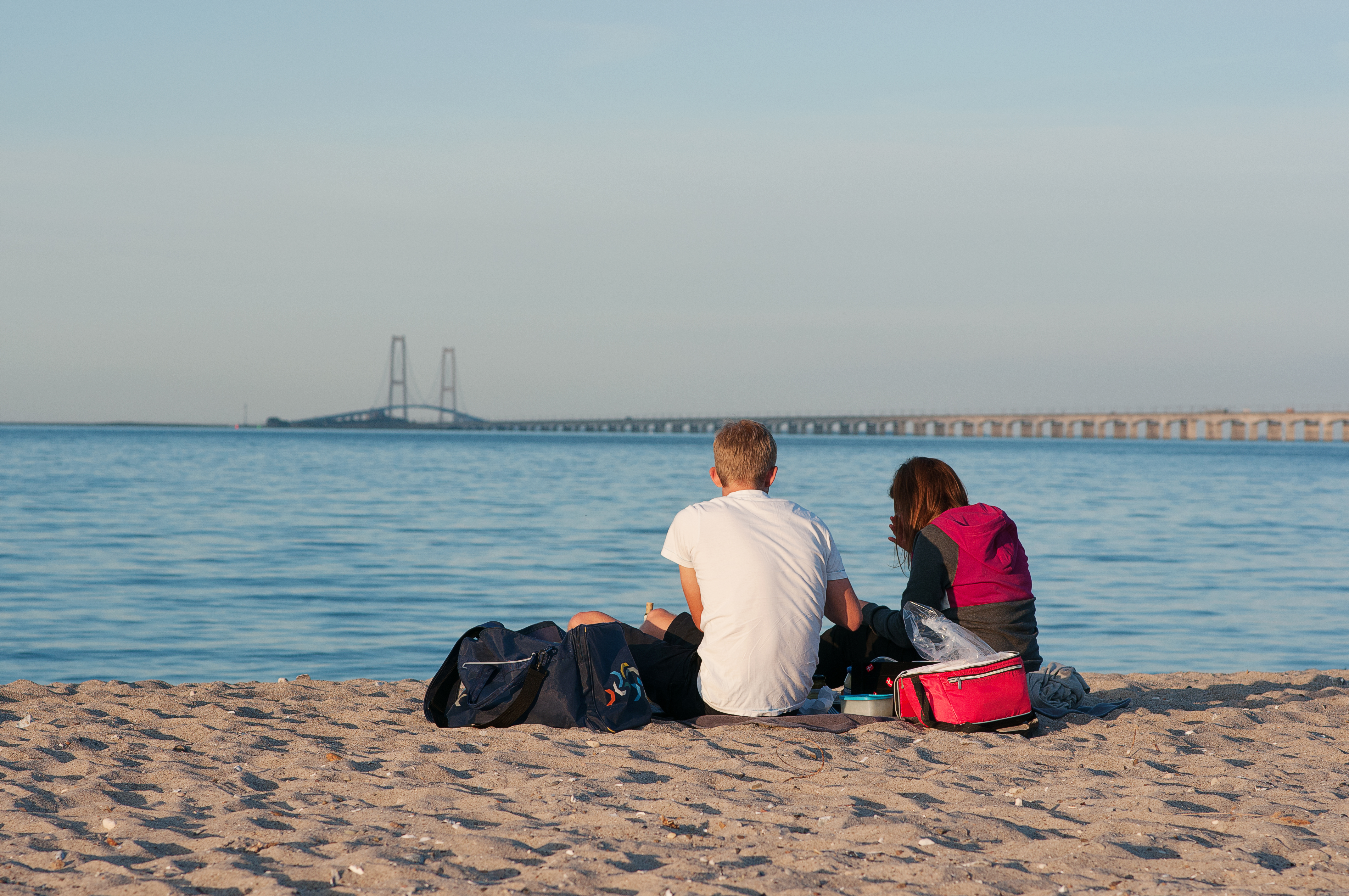 Ryggen af to unge der sidde på stranden og kigger på Storebæltsbroen