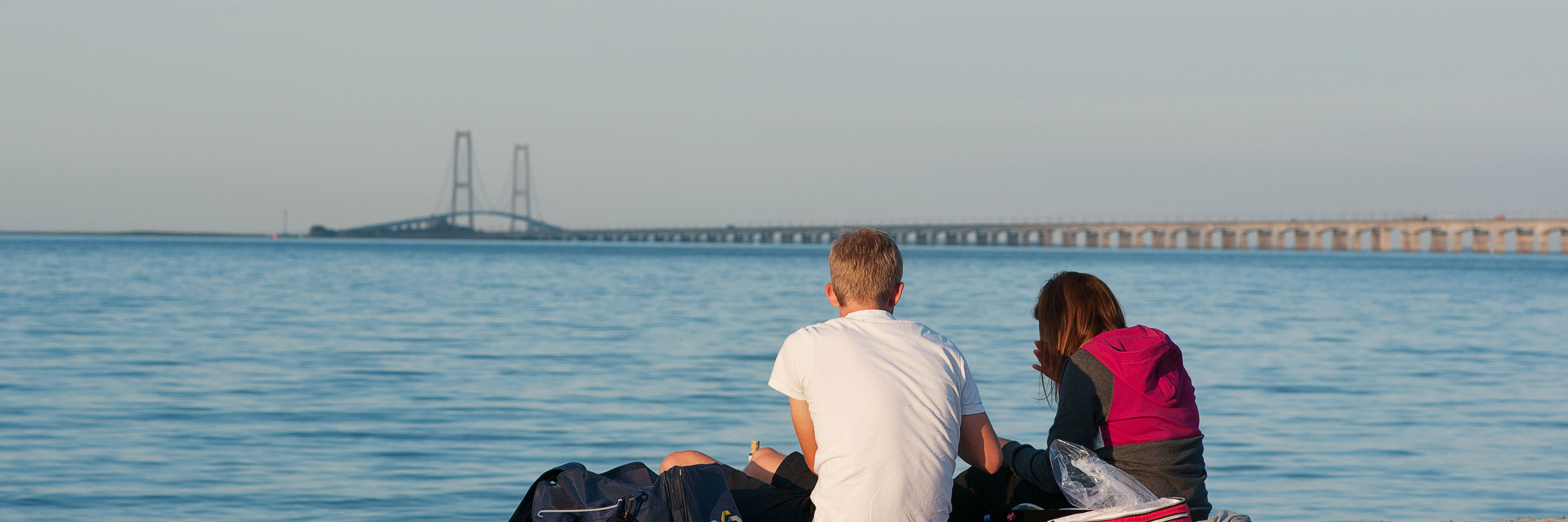 Ryggen af to unge der sidde på stranden og kigger på Storebæltsbroen