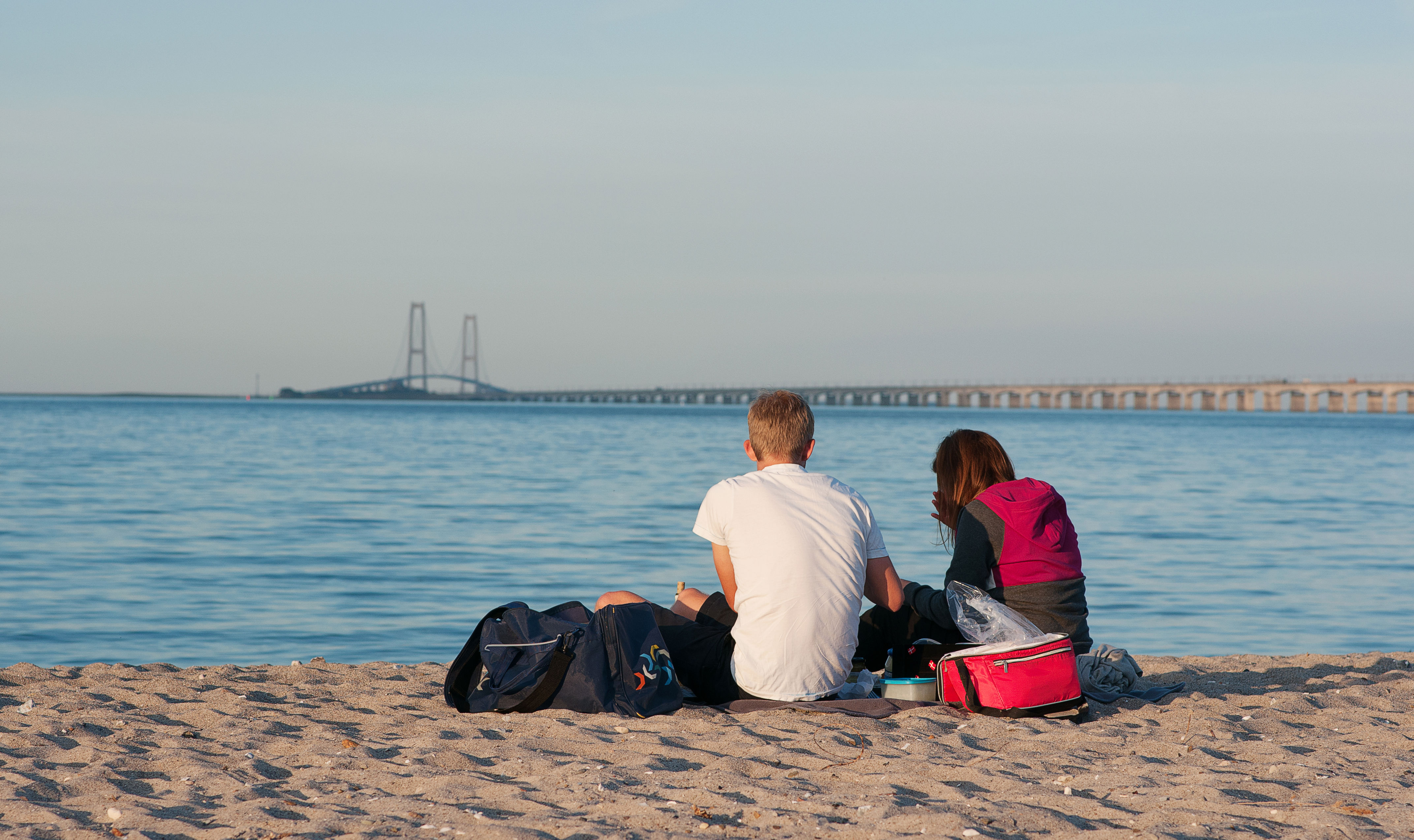 Ryggen af to unge der sidde på stranden og kigger på Storebæltsbroen